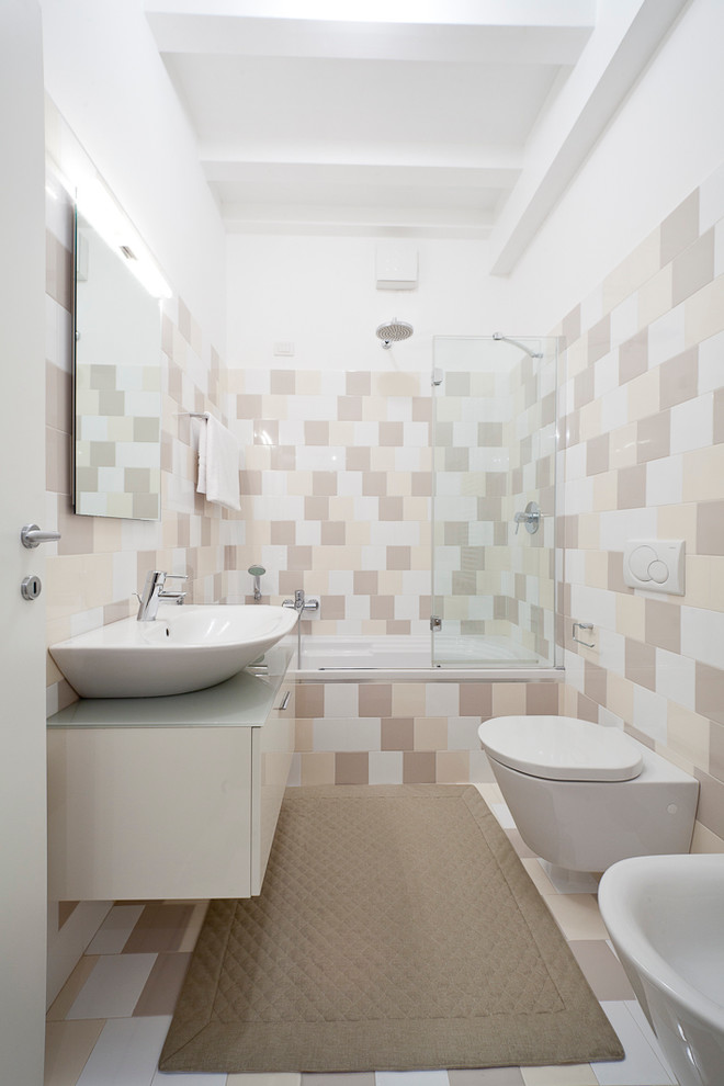 Photo of a contemporary master bathroom in Milan with a vessel sink, flat-panel cabinets, white cabinets, an alcove tub, a shower/bathtub combo, multi-coloured tile, a wall-mount toilet, ceramic tile, white walls, ceramic floors and glass benchtops.