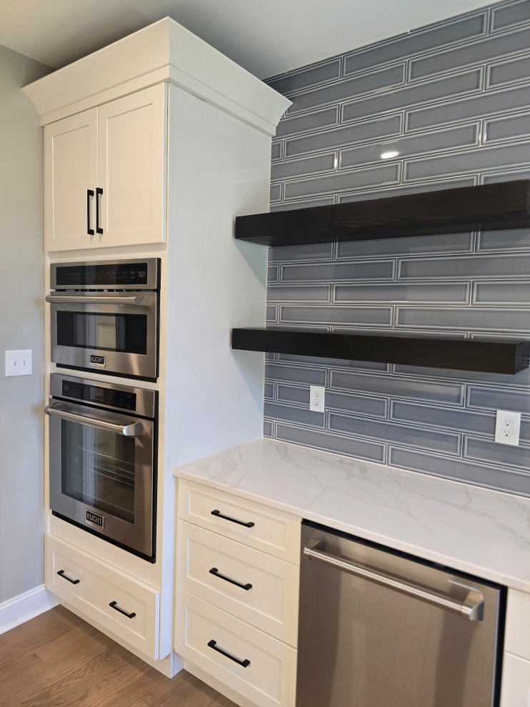 Large minimalist l-shaped medium tone wood floor and brown floor kitchen photo with shaker cabinets, white cabinets, quartz countertops, gray backsplash, subway tile backsplash, stainless steel appliances and white countertops