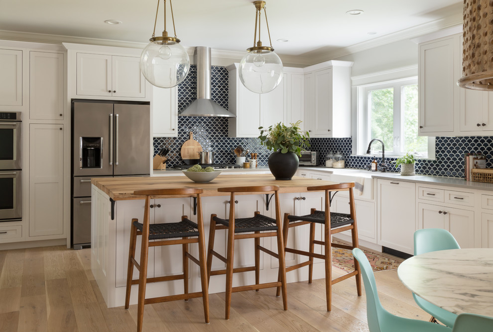 Design ideas for a beach style l-shaped eat-in kitchen in Boston with a farmhouse sink, shaker cabinets, white cabinets, blue splashback, ceramic splashback, light hardwood floors, wood benchtops, stainless steel appliances, with island and beige floor.