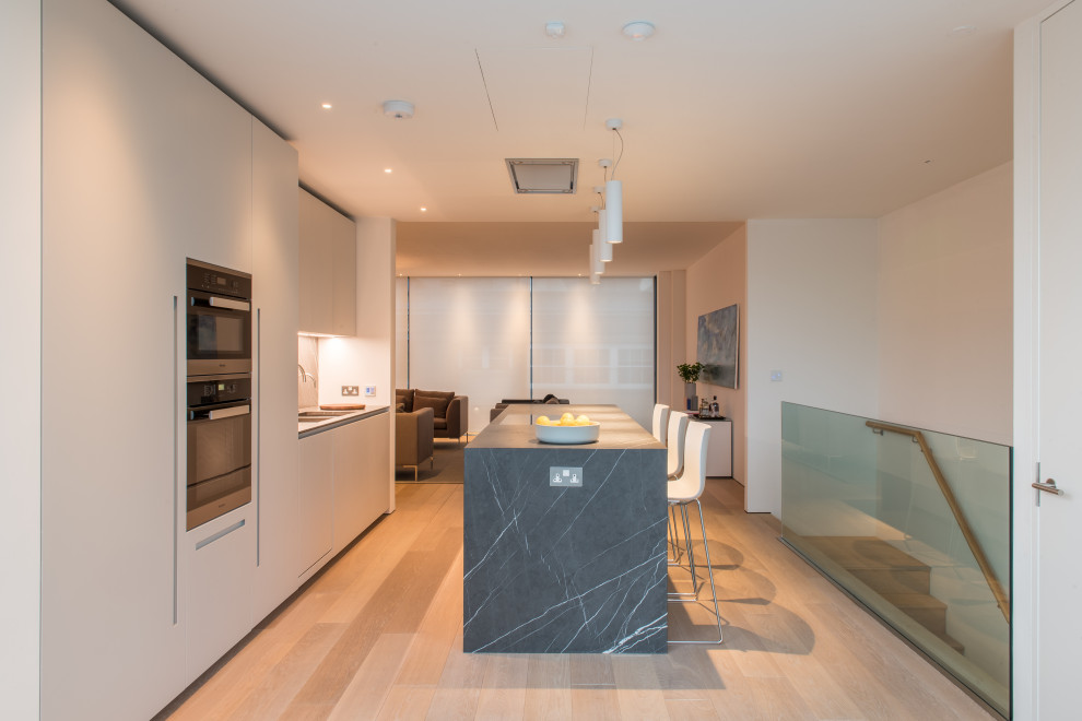 Design ideas for a contemporary single-wall kitchen/diner in London with flat-panel cabinets, white cabinets, marble worktops, grey splashback, marble splashback, stainless steel appliances, light hardwood flooring, a breakfast bar and grey worktops.