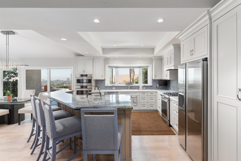 Photo of a transitional l-shaped open plan kitchen in Orange County with recessed-panel cabinets, white cabinets, blue splashback, stainless steel appliances, light hardwood floors, with island and beige floor.