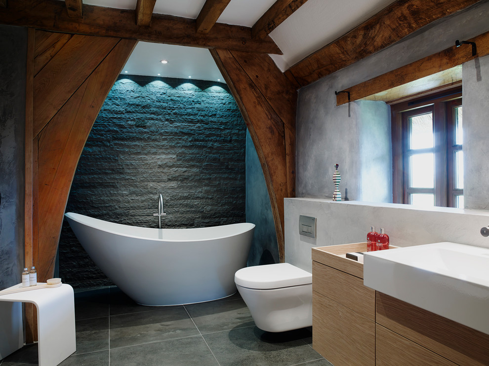 Inspiration for a mid-sized country bathroom in Devon with a wall-mount sink, a freestanding tub, an open shower, a wall-mount toilet, gray tile, stone tile, grey walls and slate floors.