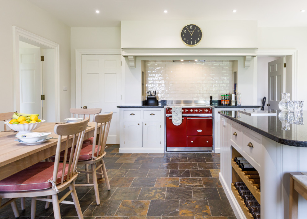 Photo of a classic kitchen/diner in Wiltshire with recessed-panel cabinets, white cabinets, white splashback, metro tiled splashback, an island, brown floors and black worktops.