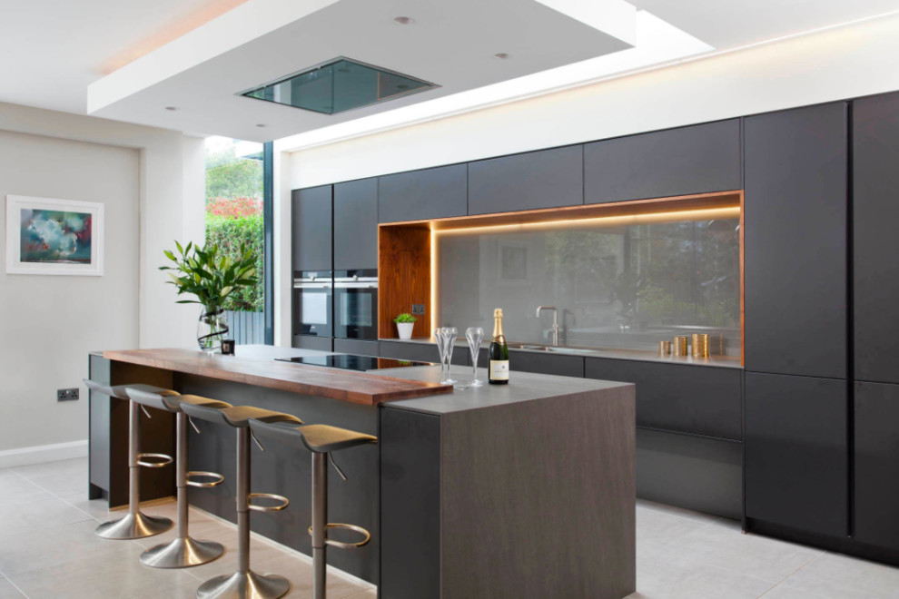 Photo of a large modern galley separate kitchen in DC Metro with a drop-in sink, flat-panel cabinets, black cabinets, wood benchtops, grey splashback, black appliances, porcelain floors, with island, beige floor, glass sheet splashback and brown benchtop.