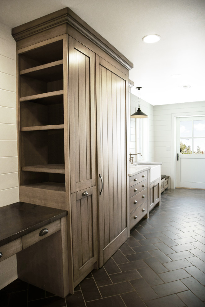 Inspiration for a mid-sized transitional single-wall dedicated laundry room in Phoenix with a farmhouse sink, shaker cabinets, medium wood cabinets, wood benchtops, grey splashback, timber splashback, white walls, limestone floors, a concealed washer and dryer, black floor, black benchtop and wood walls.