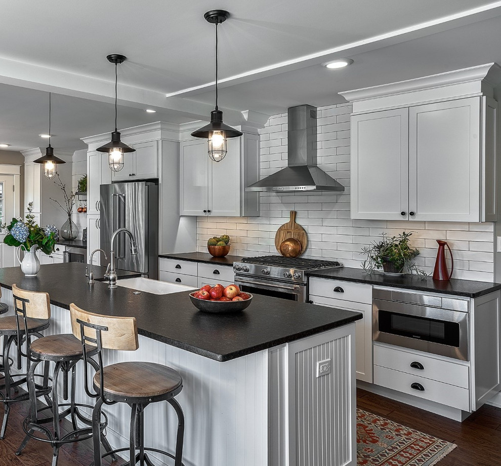 Photo of a mid-sized country galley eat-in kitchen in Chicago with a farmhouse sink, shaker cabinets, white cabinets, granite benchtops, white splashback, ceramic splashback, stainless steel appliances, medium hardwood floors, with island, brown floor and black benchtop.
