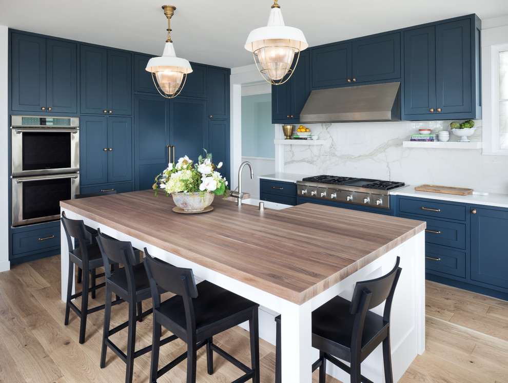 Photo of a beach style kitchen in Minneapolis with a farmhouse sink, shaker cabinets, blue cabinets, white splashback, marble splashback, stainless steel appliances, light hardwood floors, with island, beige floor and white benchtop.