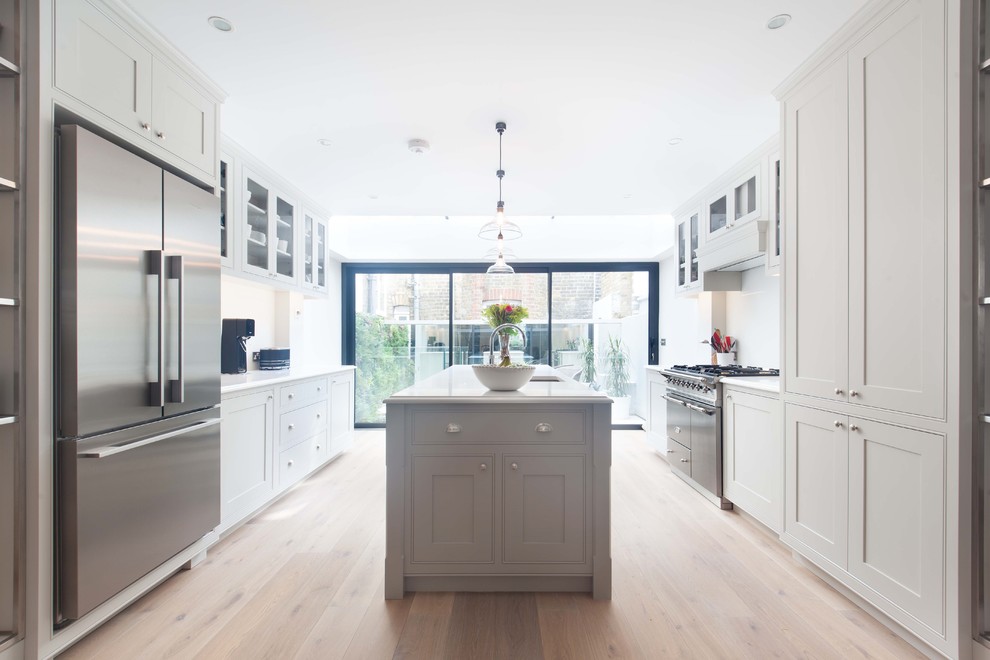 This is an example of a mid-sized transitional galley eat-in kitchen in London with shaker cabinets, with island, an undermount sink, beige cabinets, white splashback, stainless steel appliances, light hardwood floors, beige floor and white benchtop.