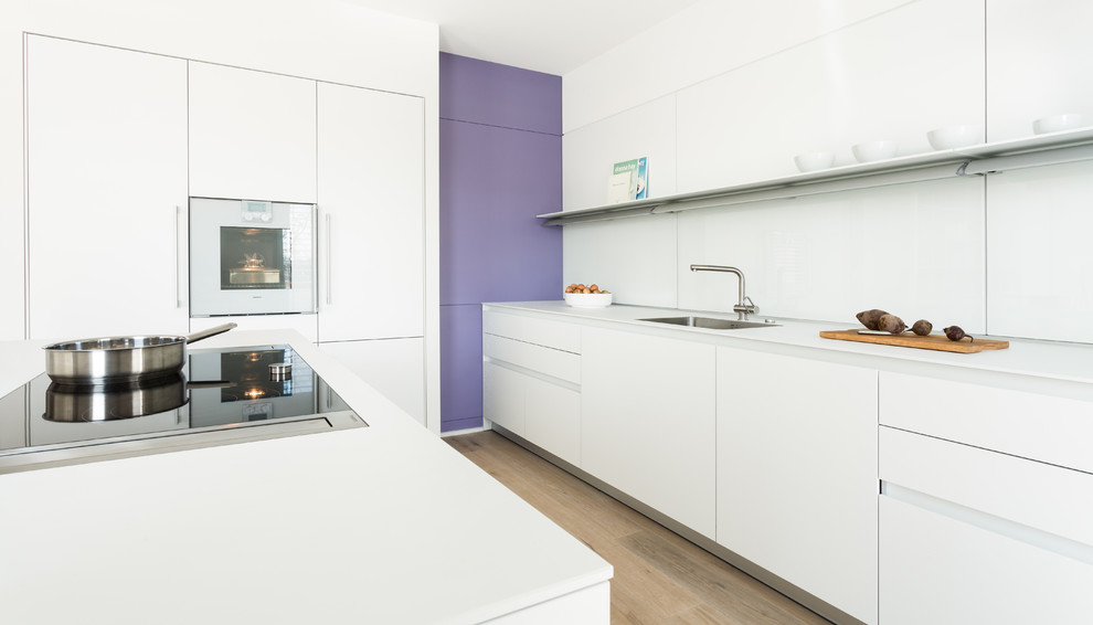 Design ideas for a contemporary single-wall kitchen in Stuttgart with a drop-in sink, flat-panel cabinets, white cabinets, white splashback, glass sheet splashback, medium hardwood floors, with island and brown floor.