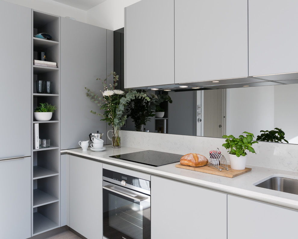 Mid-sized contemporary l-shaped eat-in kitchen in Gloucestershire with an integrated sink, flat-panel cabinets, grey cabinets, solid surface benchtops, grey splashback, mirror splashback, black appliances, laminate floors, no island, brown floor and white benchtop.