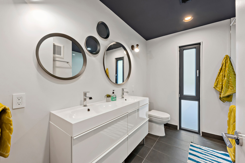 This is an example of a mid-sized contemporary 3/4 bathroom in Charlotte with flat-panel cabinets, white cabinets, an alcove tub, a shower/bathtub combo, a two-piece toilet, white tile, porcelain tile, white walls, porcelain floors, an integrated sink, engineered quartz benchtops, black floor, an open shower and white benchtops.