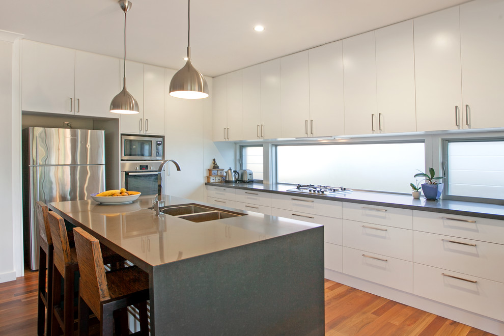 Photo of a contemporary u-shaped kitchen in Central Coast with an undermount sink, flat-panel cabinets, white cabinets, window splashback, stainless steel appliances, medium hardwood floors, with island, brown floor and grey benchtop.