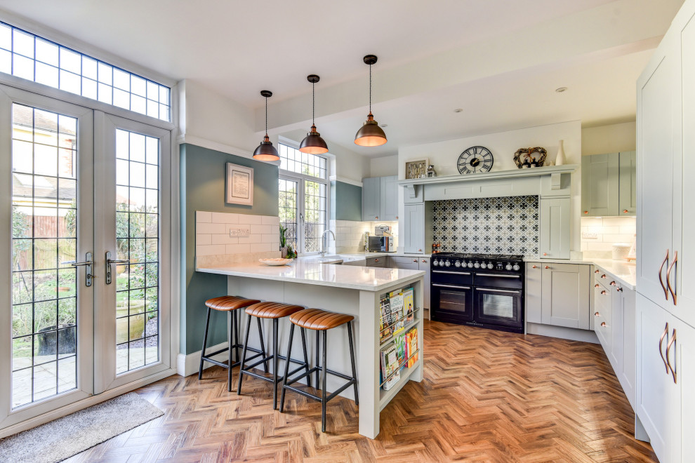 Inspiration for a large classic grey and teal u-shaped open plan kitchen in Sussex with a belfast sink, shaker cabinets, blue cabinets, quartz worktops, white splashback, metro tiled splashback, black appliances, medium hardwood flooring, a breakfast bar, brown floors, white worktops and a chimney breast.