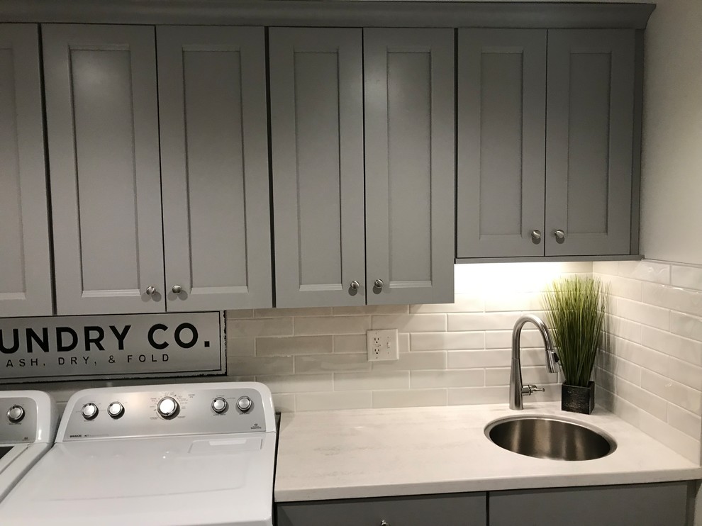 This is an example of a mid-sized contemporary single-wall utility room in New York with an undermount sink, recessed-panel cabinets, grey cabinets, solid surface benchtops, grey walls, a side-by-side washer and dryer and white benchtop.