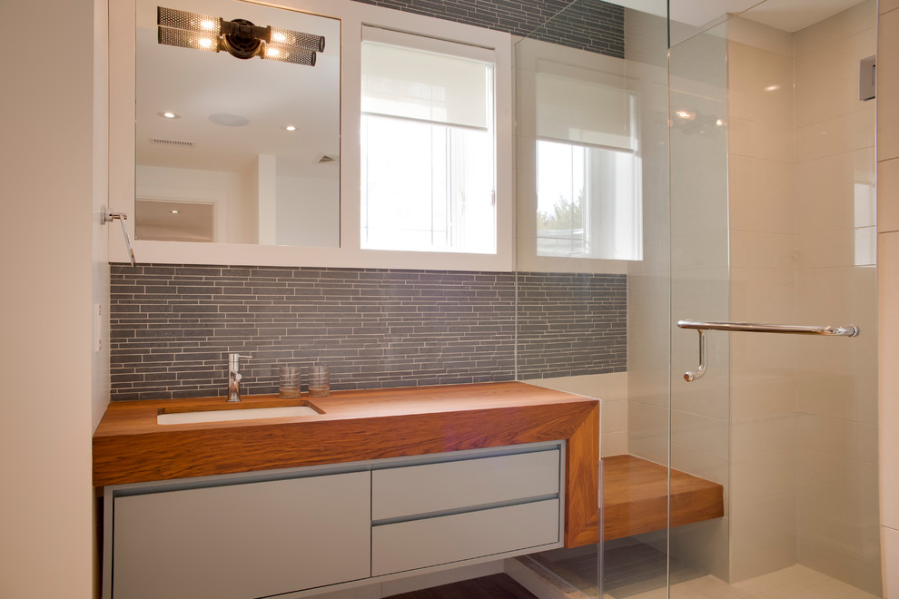 Mid-sized modern master bathroom with a freestanding tub, a one-piece toilet, an undermount sink, flat-panel cabinets, grey cabinets, an alcove shower, gray tile, glass tile, white walls, light hardwood floors, wood benchtops, brown floor and a hinged shower door.