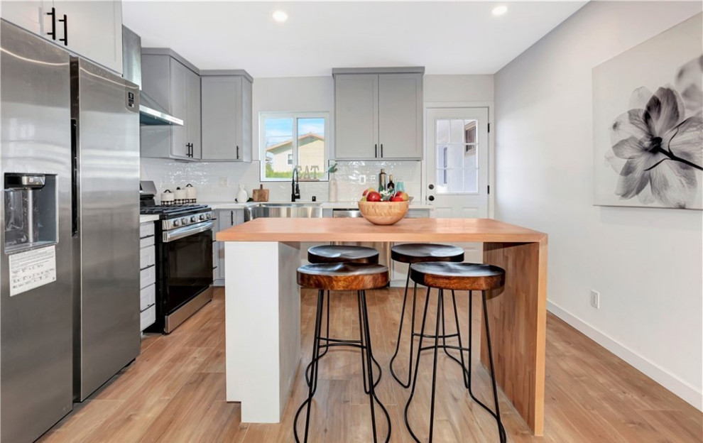 This is an example of a contemporary l-shaped kitchen in Los Angeles with a built-in sink, recessed-panel cabinets, grey cabinets, composite countertops, white splashback, ceramic splashback, stainless steel appliances, medium hardwood flooring, an island, brown floors and white worktops.