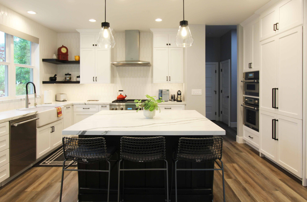 Example of a large transitional l-shaped vinyl floor and brown floor open concept kitchen design with a farmhouse sink, shaker cabinets, white cabinets, quartz countertops, white backsplash, ceramic backsplash, stainless steel appliances, an island and white countertops