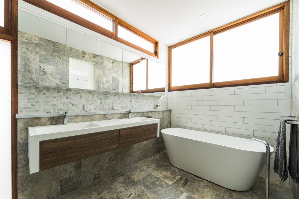 Inspiration for a large contemporary master bathroom in Brisbane with flat-panel cabinets, medium wood cabinets, a freestanding tub, an open shower, a wall-mount toilet, multi-coloured tile, travertine, white walls, travertine floors, an integrated sink, solid surface benchtops, brown floor and an open shower.
