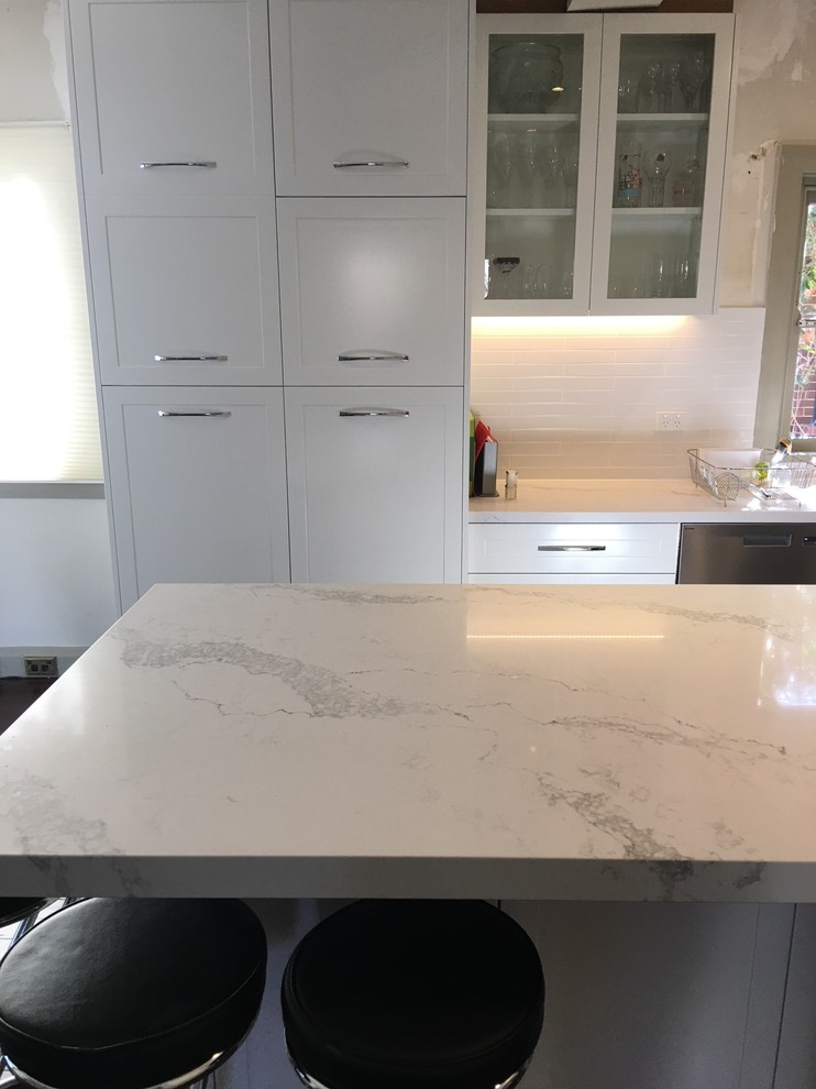 This is an example of a mid-sized transitional l-shaped eat-in kitchen in Sydney with a drop-in sink, recessed-panel cabinets, white cabinets, quartz benchtops, white splashback, subway tile splashback, stainless steel appliances, medium hardwood floors, with island and brown floor.
