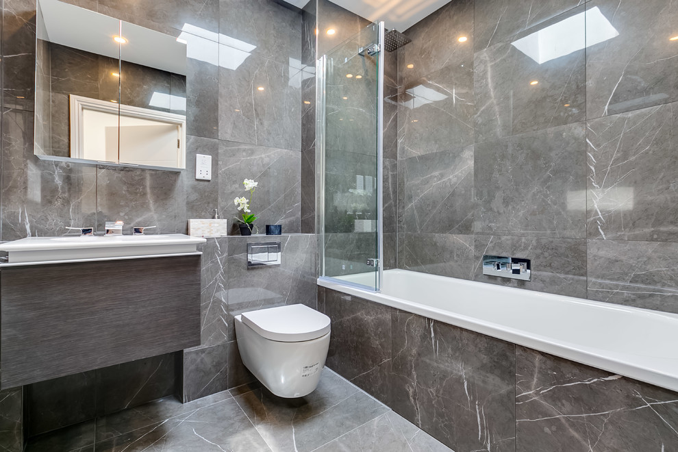 Mid-sized contemporary kids bathroom in London with a drop-in tub, a shower/bathtub combo, a wall-mount toilet, grey walls, a drop-in sink, grey floor, flat-panel cabinets, grey cabinets, gray tile and an open shower.