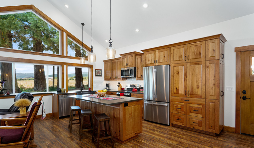 Example of a large mountain style medium tone wood floor, brown floor and vaulted ceiling kitchen design with a farmhouse sink, raised-panel cabinets, medium tone wood cabinets, concrete countertops, white backsplash, ceramic backsplash, stainless steel appliances, an island and gray countertops