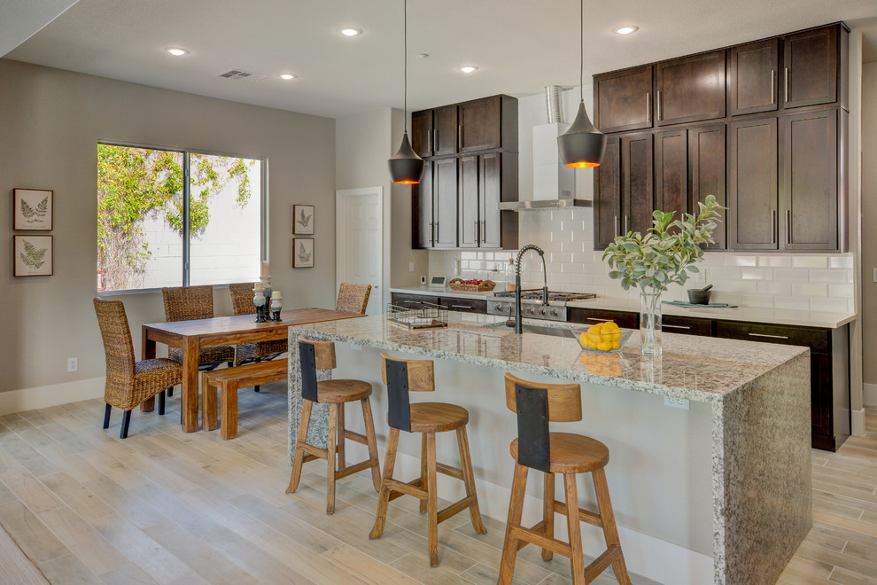 This is an example of a mid-sized transitional l-shaped open plan kitchen in Las Vegas with a farmhouse sink, shaker cabinets, dark wood cabinets, granite benchtops, white splashback, porcelain splashback, stainless steel appliances, porcelain floors, with island and grey floor.