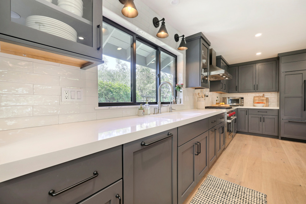 Mid-sized transitional l-shaped laminate floor open concept kitchen photo with a double-bowl sink, flat-panel cabinets, gray cabinets, quartz countertops, white backsplash, ceramic backsplash, stainless steel appliances, an island and white countertops