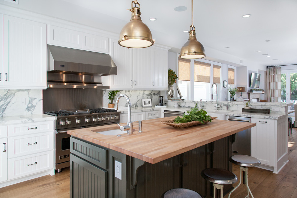 Photo of a beach style l-shaped open plan kitchen in Orange County with a farmhouse sink, white cabinets, wood benchtops, white splashback and stainless steel appliances.