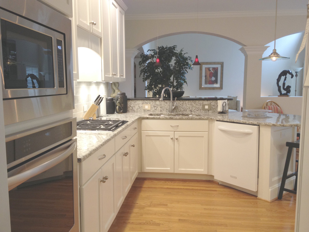 Inspiration for a mid-sized transitional l-shaped light wood floor eat-in kitchen remodel with an undermount sink, recessed-panel cabinets, white cabinets, granite countertops, white backsplash, stone tile backsplash, white appliances and a peninsula