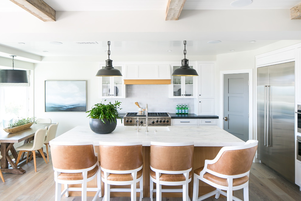 Inspiration for a beach style eat-in kitchen in Los Angeles with white cabinets, white splashback, subway tile splashback, stainless steel appliances, light hardwood floors, with island, black benchtop and shaker cabinets.