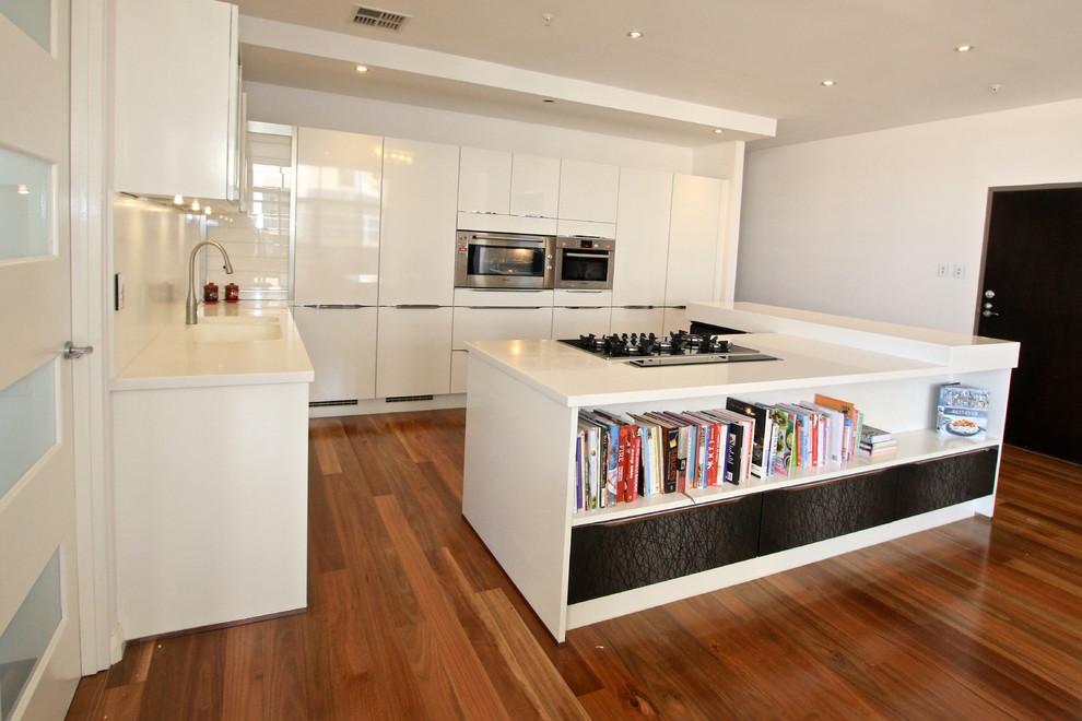 Design ideas for a large modern l-shaped open plan kitchen in Perth with an integrated sink, flat-panel cabinets, white cabinets, solid surface benchtops, white splashback, glass sheet splashback, black appliances, medium hardwood floors and with island.