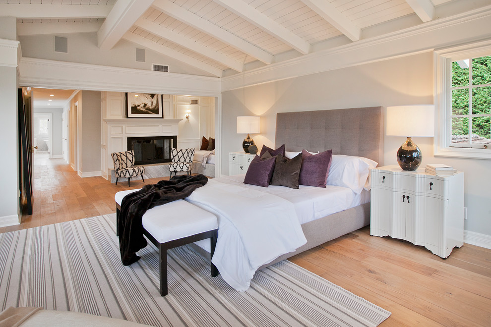 Photo of a large transitional master bedroom in Los Angeles with grey walls, light hardwood floors, a standard fireplace, a wood fireplace surround and beige floor.