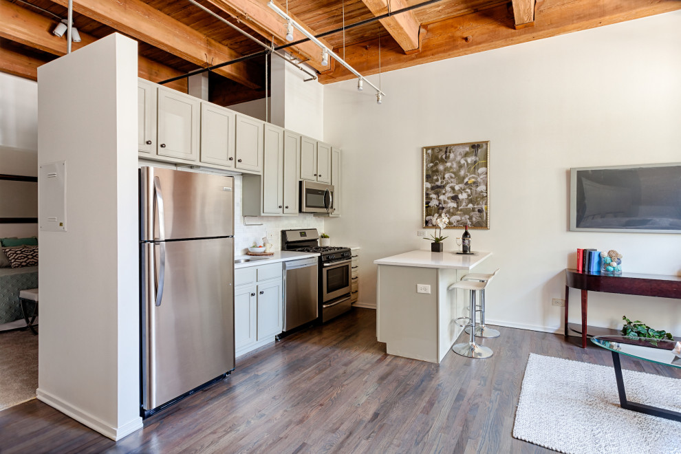 Photo of an industrial galley open plan kitchen in Chicago with shaker cabinets, grey cabinets, stainless steel appliances, dark hardwood floors, a peninsula, brown floor, white benchtop, exposed beam and wood.