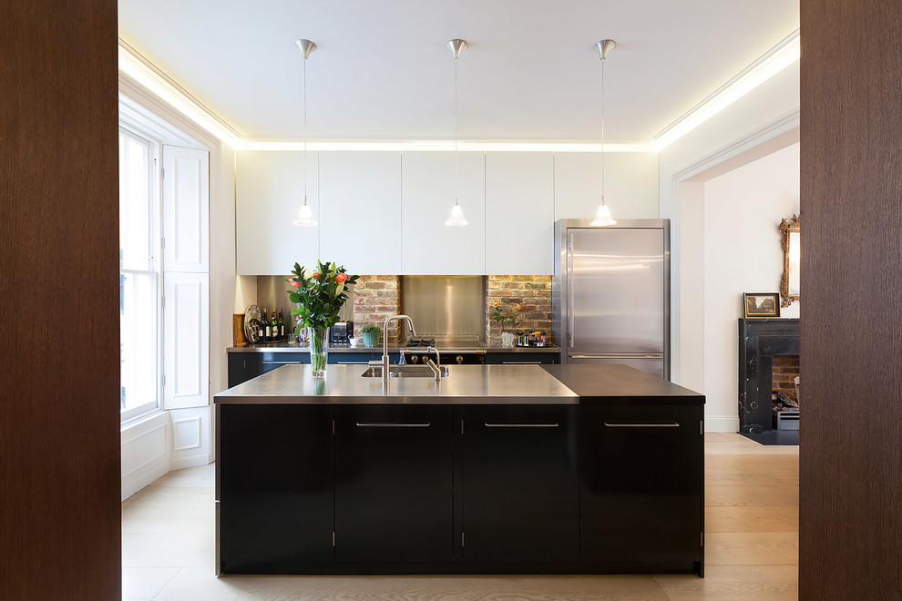 This is an example of a mid-sized contemporary galley separate kitchen in London with a double-bowl sink, flat-panel cabinets, brick splashback, stainless steel appliances, light hardwood floors, with island, brown floor, black cabinets and stainless steel benchtops.