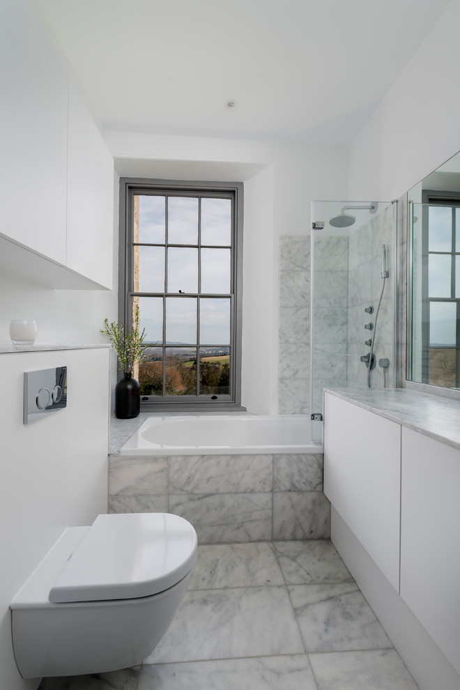 Photo of a mid-sized contemporary master bathroom in Devon with flat-panel cabinets, white cabinets, a drop-in tub, a curbless shower, a wall-mount toilet, white tile, stone slab, white walls, marble floors, a drop-in sink, marble benchtops, white floor and an open shower.