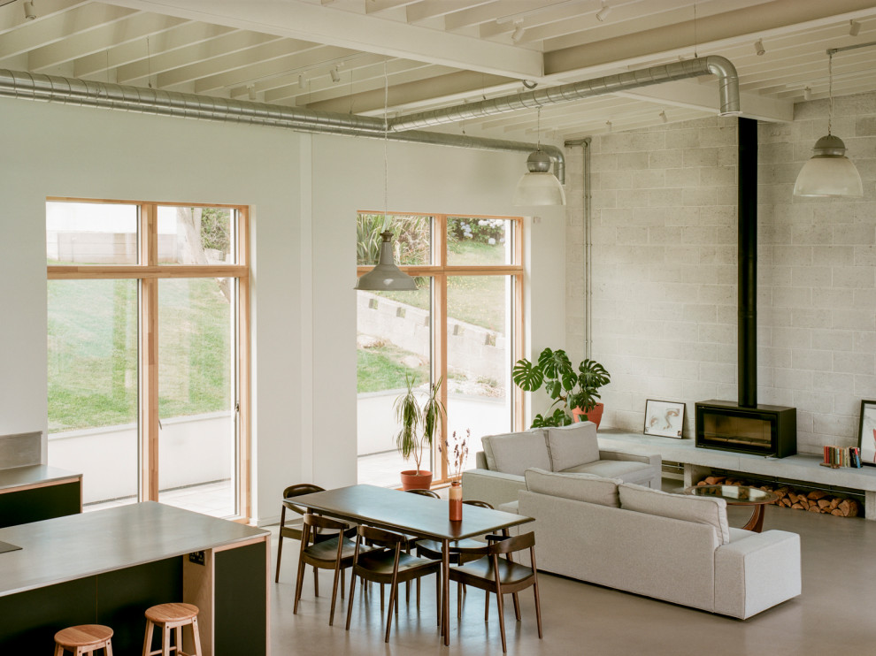 Large open plan dining room in Cornwall with White Walls, Concrete Flooring, a Wood Burning Stove, a Metal Fireplace Surround, Grey Floors, Exposed Beams, Brick Walls and a Feature Wall.