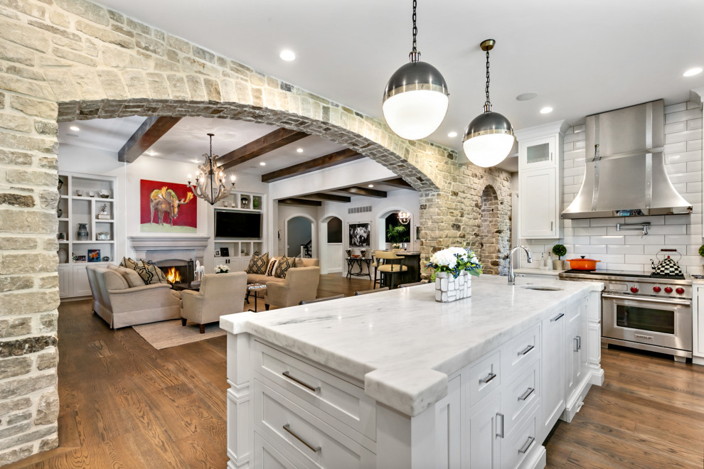 Elegant l-shaped medium tone wood floor and brown floor kitchen photo in St Louis with a farmhouse sink, shaker cabinets, white cabinets, white backsplash, subway tile backsplash, stainless steel appliances, an island and white countertops
