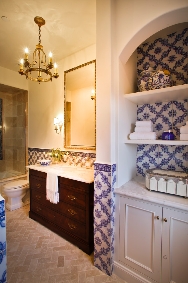 Photo of a mid-sized mediterranean kids bathroom in Los Angeles with recessed-panel cabinets, dark wood cabinets, an undermount tub, a shower/bathtub combo, blue tile, ceramic tile, white walls, limestone floors, an undermount sink, marble benchtops, beige floor, a hinged shower door and white benchtops.