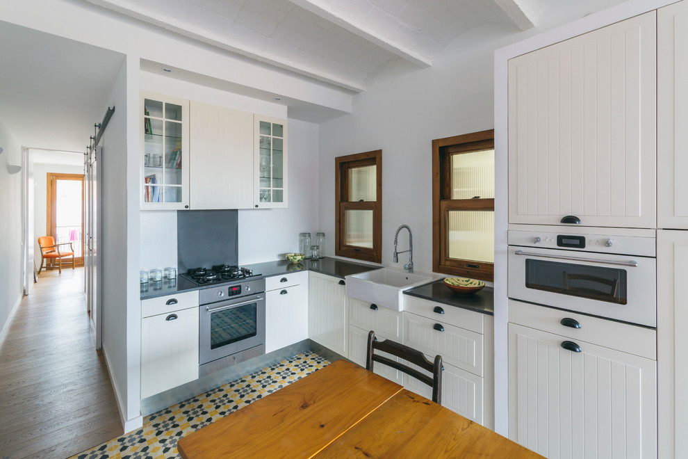 Mid-sized scandinavian l-shaped eat-in kitchen in Barcelona with a farmhouse sink, raised-panel cabinets, white cabinets, white splashback, stainless steel appliances, ceramic floors and no island.