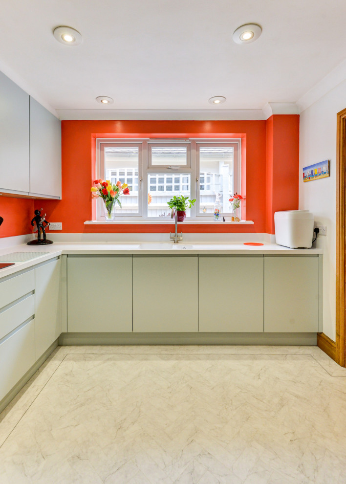 This is an example of a medium sized beach style grey and white l-shaped enclosed kitchen in Sussex with a built-in sink, flat-panel cabinets, green cabinets, composite countertops, orange splashback, glass sheet splashback, black appliances, lino flooring, a breakfast bar, white floors, white worktops and feature lighting.