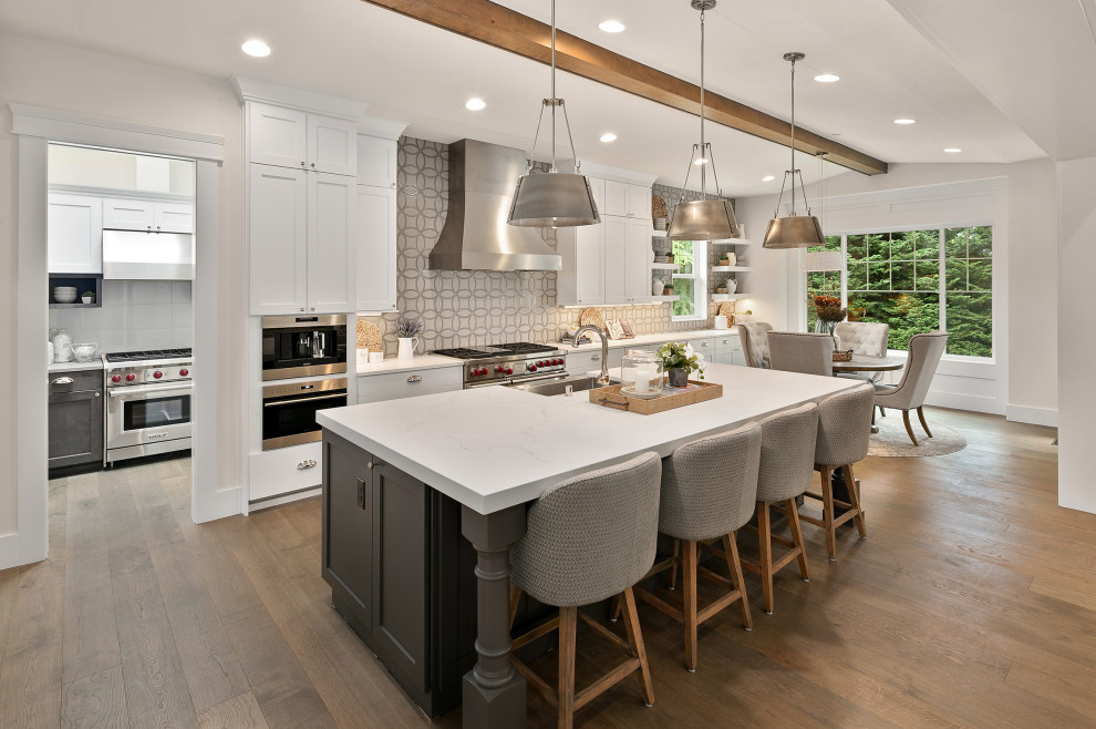 Photo of a large transitional single-wall eat-in kitchen in Seattle with a farmhouse sink, shaker cabinets, white cabinets, quartzite benchtops, grey splashback, porcelain splashback, stainless steel appliances, medium hardwood floors, with island, brown floor, white benchtop and exposed beam.