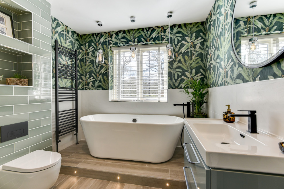 This is an example of a medium sized grey and white family bathroom in Sussex with flat-panel cabinets, blue cabinets, a freestanding bath, a walk-in shower, a one-piece toilet, green tiles, ceramic tiles, green walls, light hardwood flooring, a built-in sink, brown floors, a hinged door, white worktops, feature lighting, a single sink, a floating vanity unit and wallpapered walls.