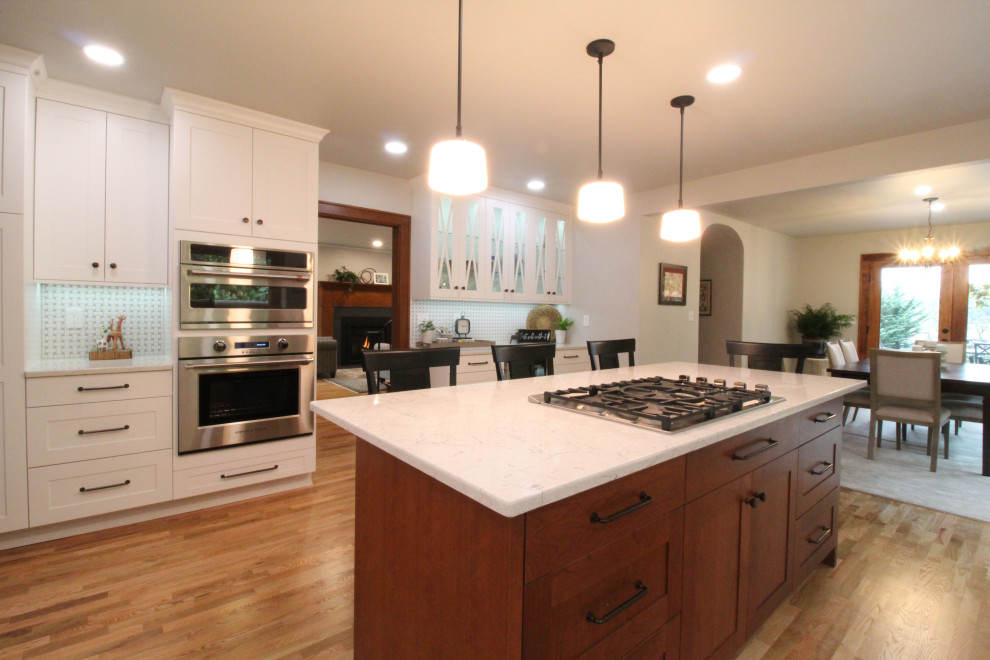 Photo of a medium sized classic l-shaped kitchen/diner in Seattle with a belfast sink, recessed-panel cabinets, white cabinets, engineered stone countertops, white splashback, ceramic splashback, stainless steel appliances, light hardwood flooring, an island, brown floors and white worktops.