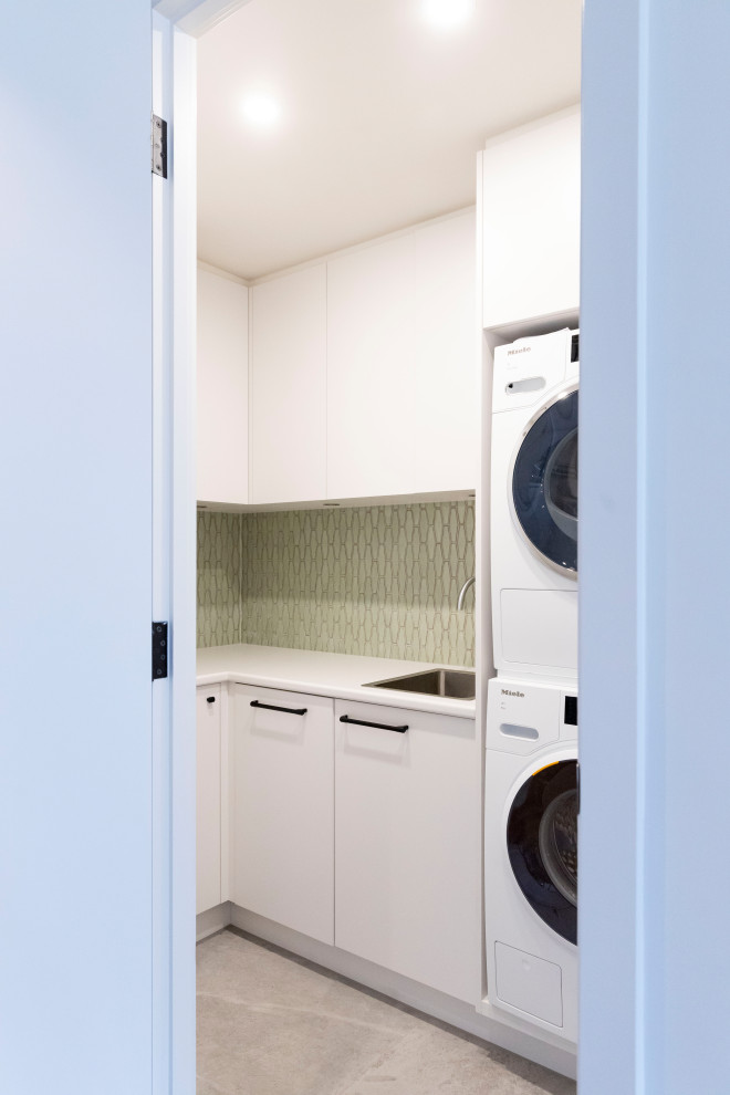 Mid-sized contemporary l-shaped dedicated laundry room in Melbourne with a single-bowl sink, flat-panel cabinets, white cabinets, laminate benchtops, green splashback, ceramic splashback, white walls, a stacked washer and dryer, white benchtop, porcelain floors and grey floor.