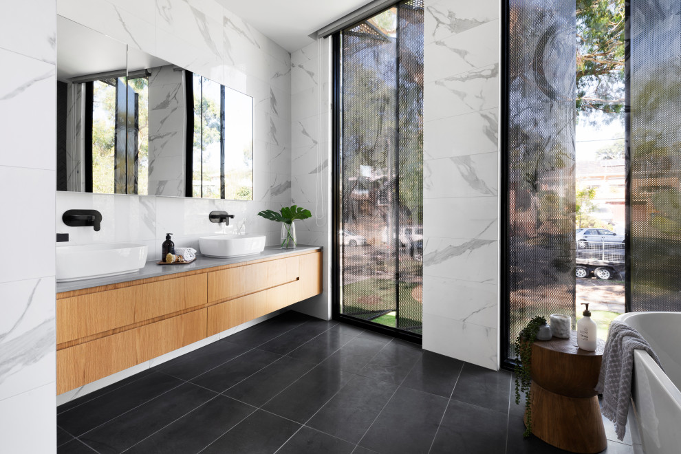 Inspiration for a large contemporary master bathroom in Melbourne with flat-panel cabinets, medium wood cabinets, a freestanding tub, white tile, a vessel sink, black floor, grey benchtops, a double vanity, a floating vanity, an alcove shower, a one-piece toilet, porcelain tile, white walls, porcelain floors, engineered quartz benchtops, a hinged shower door and a niche.