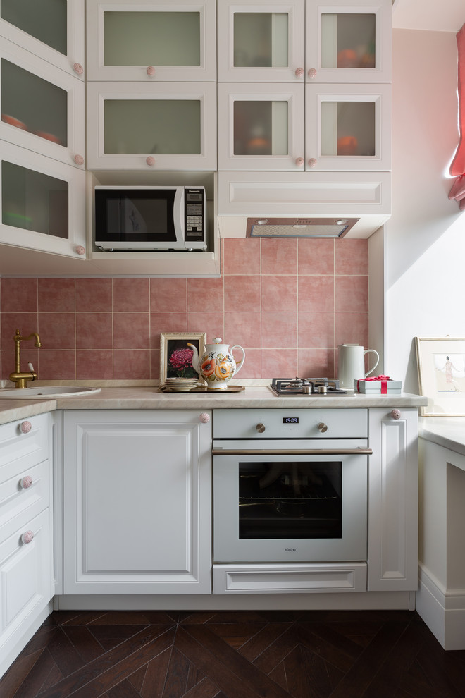 This is an example of a small contemporary l-shaped kitchen in Saint Petersburg with a drop-in sink, glass-front cabinets, white cabinets, pink splashback, white appliances, dark hardwood floors, brown floor and beige benchtop.