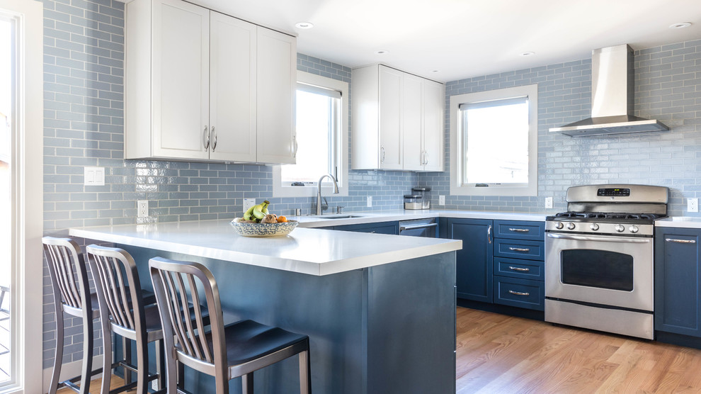 This is an example of a transitional u-shaped kitchen in San Francisco with an undermount sink, shaker cabinets, blue cabinets, blue splashback, subway tile splashback, stainless steel appliances, medium hardwood floors, a peninsula, brown floor and white benchtop.