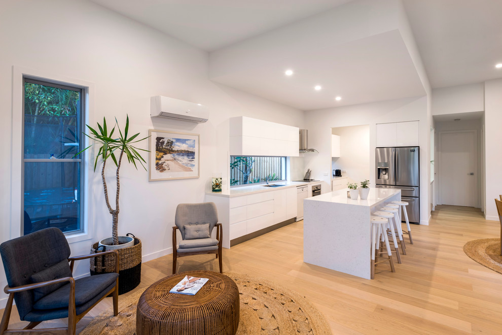 Photo of a mid-sized tropical galley open plan kitchen in Gold Coast - Tweed with a double-bowl sink, flat-panel cabinets, white cabinets, quartz benchtops, white splashback, glass sheet splashback, stainless steel appliances, light hardwood floors, with island, brown floor and white benchtop.