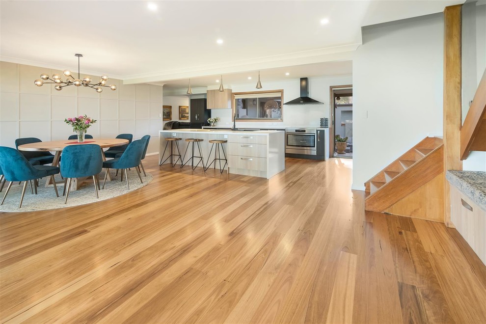 Photo of a large contemporary single-wall eat-in kitchen in Sydney with a farmhouse sink, flat-panel cabinets, beige cabinets, granite benchtops, white splashback, black appliances, light hardwood floors, with island, brown floor and white benchtop.
