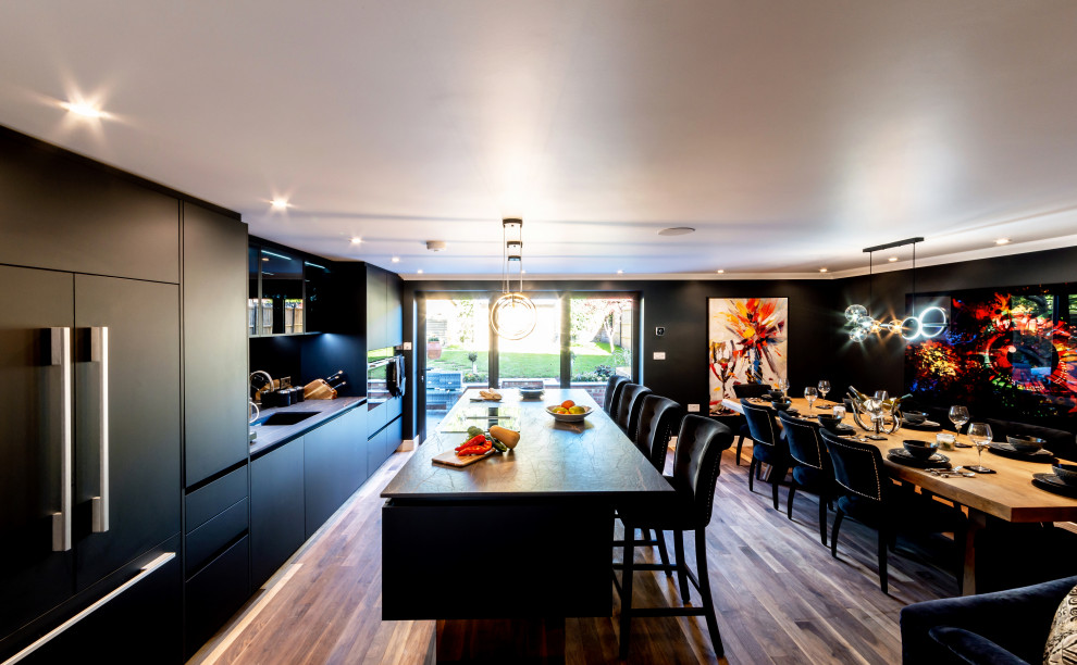 Inspiration for a medium sized contemporary grey and black kitchen/diner in Surrey with a built-in sink, flat-panel cabinets, black cabinets, granite worktops, black splashback, glass sheet splashback, black appliances, medium hardwood flooring, an island, brown floors, grey worktops and feature lighting.